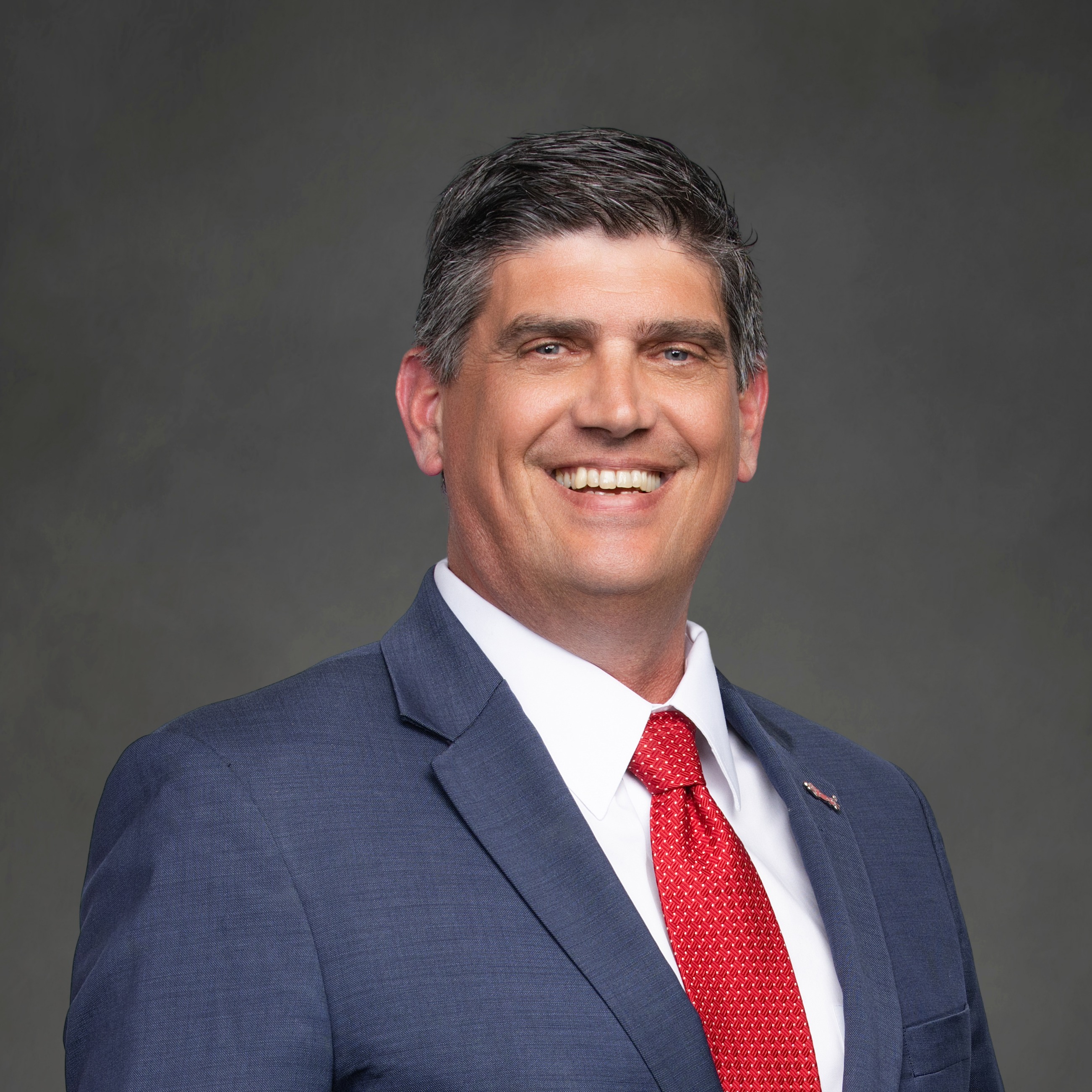 A man in a navy blue suit with a red tie and white shirt smiles at the camera.