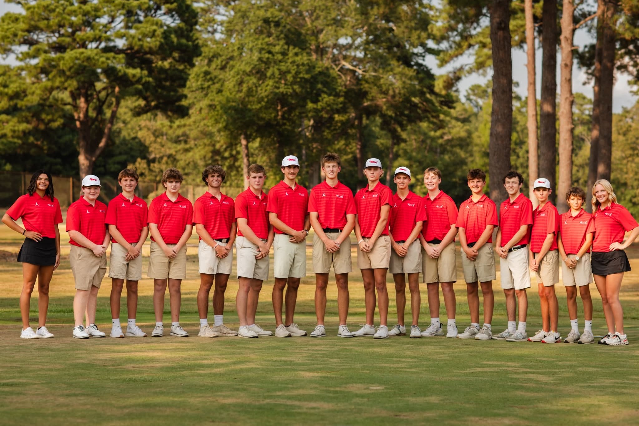 A group of students in red shirts and white shorts pose for a photo in a line on a golf course.