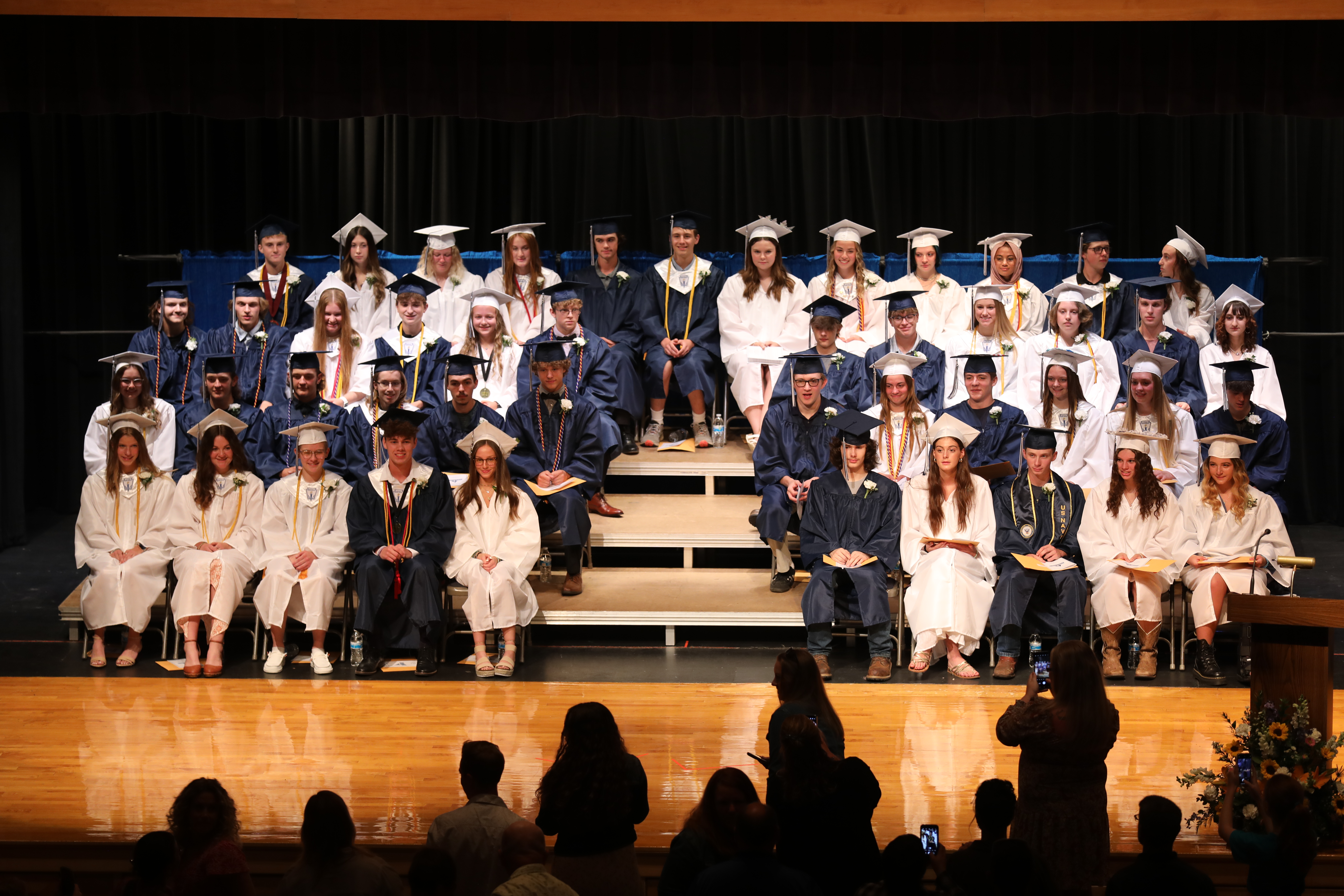 The Class of 2023 assembled on the stage at graduation. Some students are wearing white caps and gowns and some are wearing blue caps and gowns
