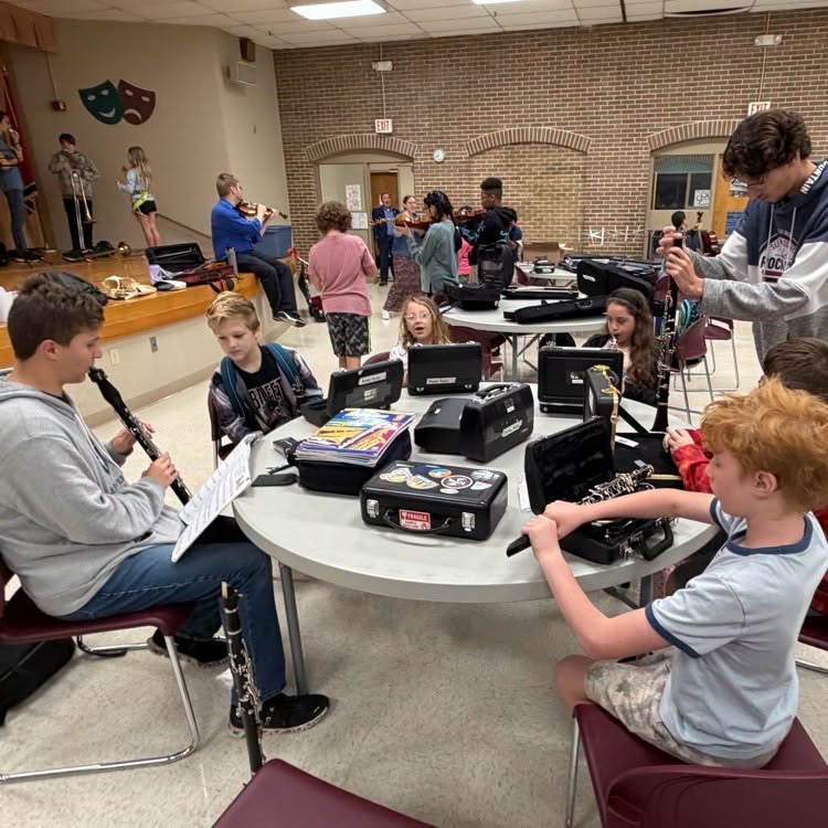 Several children play musical instruments at a table in a large room with an audience.