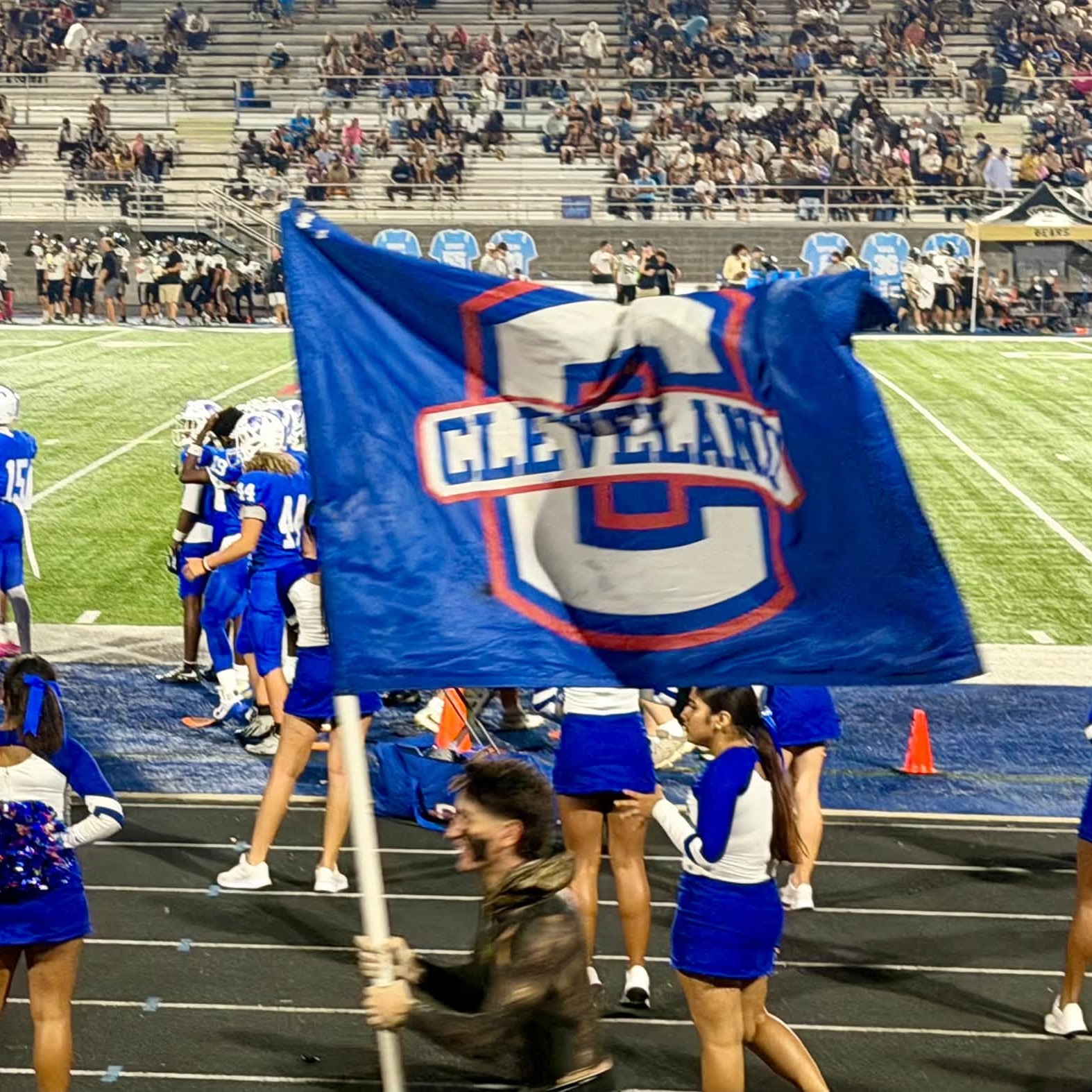 Cheerleaders carry a blue flag for Cleveland, marching on a field with players in blue. Audience in bleachers.