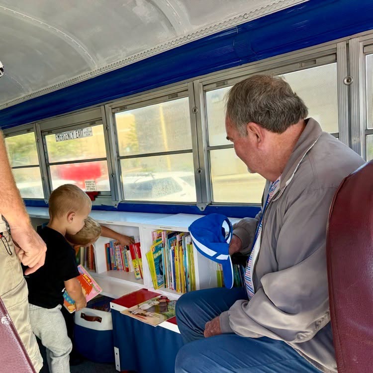 Man and child seated inside a bus, picking out books from a shelf. Another person stands nearby.