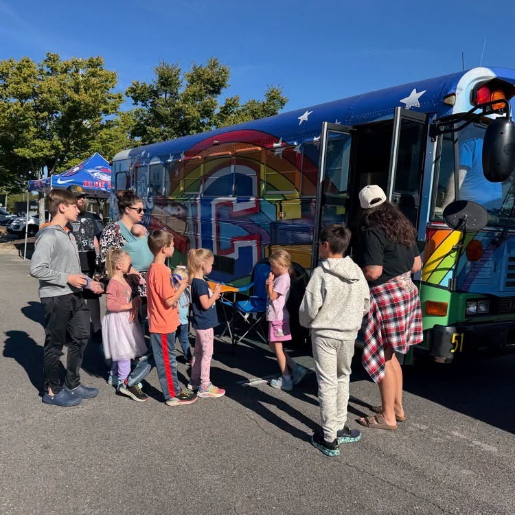 A group of people, including children, line up to enter a colorful bus with a rainbow design.