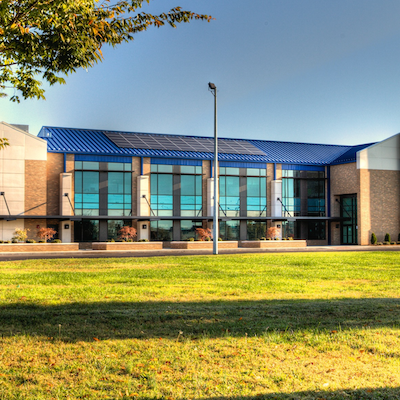 A modern building with a blue roof and solar panels. Glass windows and a streetlight are visible. Green lawn in front.