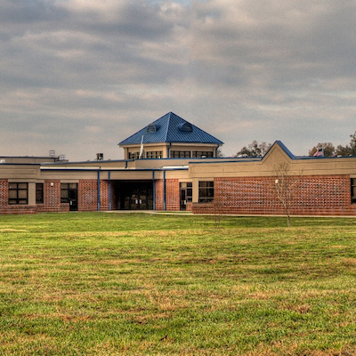 A school building with a blue roof and brick walls stands in a large, open grassy field.