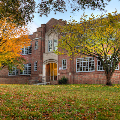 A large red brick building with arched windows, an arched entrance, and two trees in front. Leaves are on the ground.