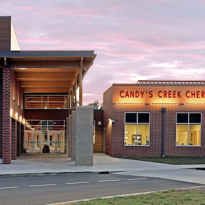 Exterior of a modern building with brick and glass elements. A sign reads "Candy's Creek." The sky shows hues of pink and blue.