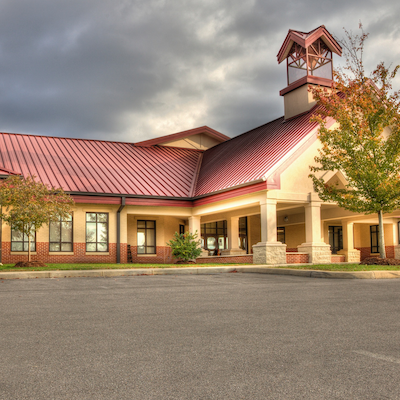 A school with a red roof, bell tower, and front entrance. Trees and plants are in front. Cloudy sky.