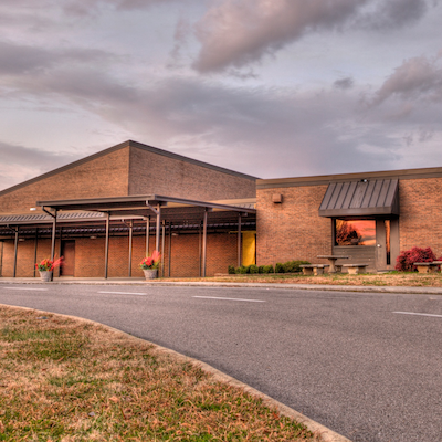 Building with gray roof and awning. Two potted plants in front. Empty road with grassy sides.