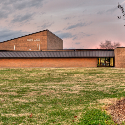 A two-story building with a gray roof stands in a wide, grassy area. The sky above is partly cloudy.