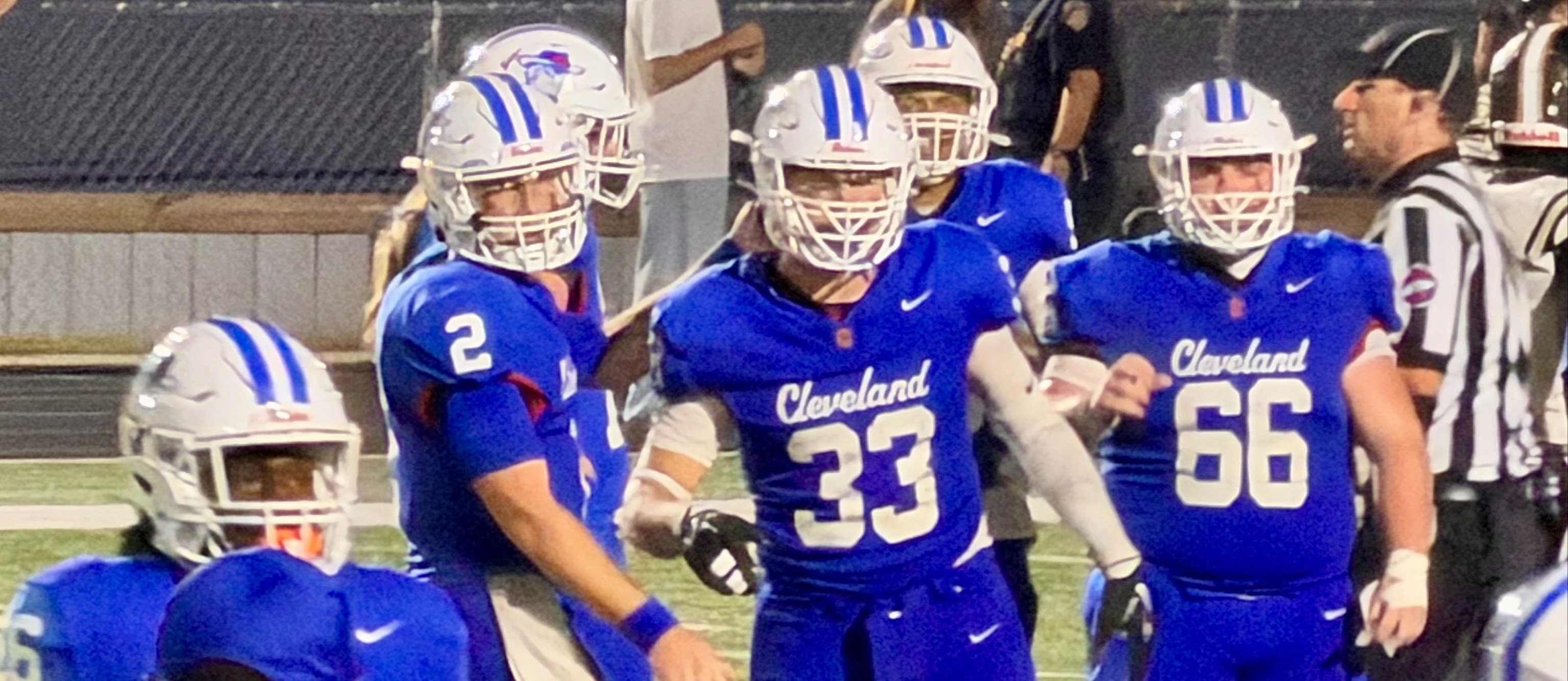 American football players in blue uniforms and helmets gather on a field, with numbers 33, 2, 66, and 5 visible.
