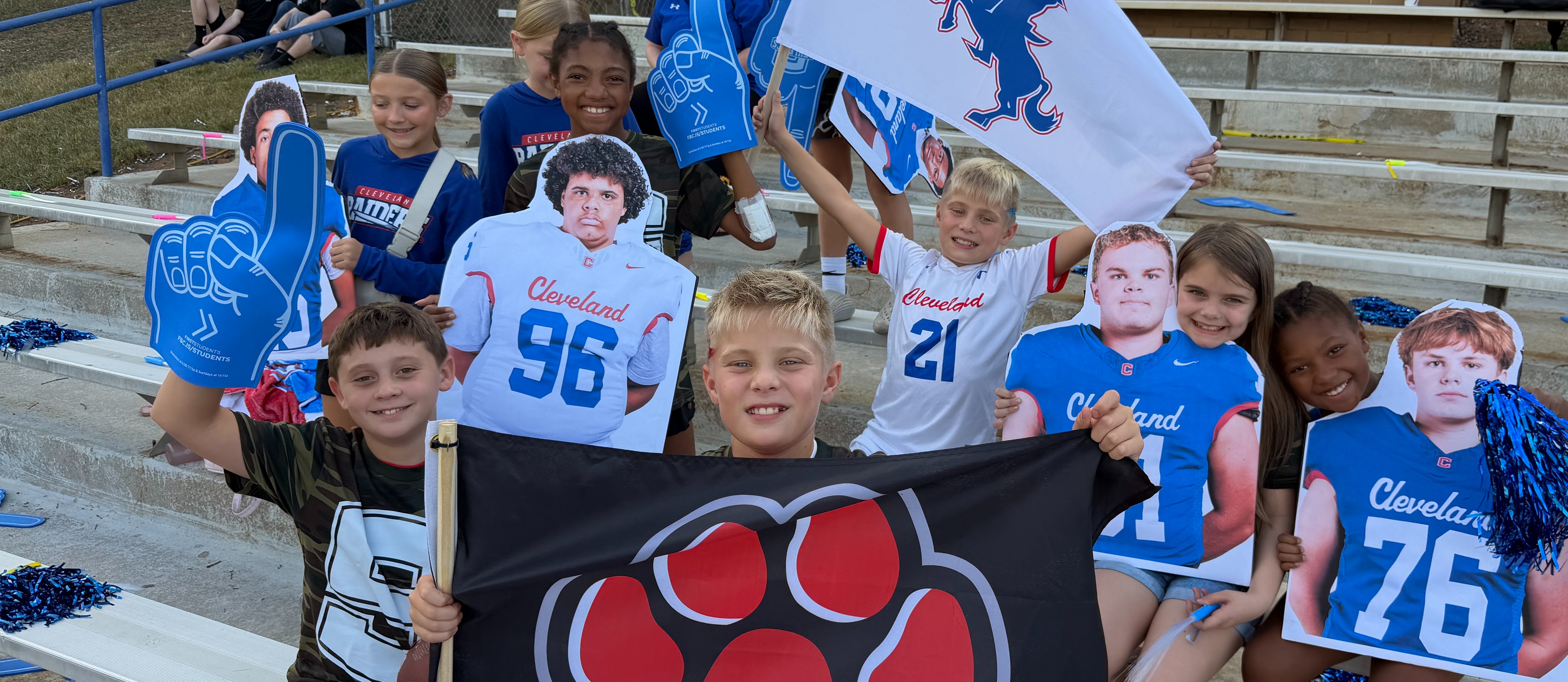 Children and adults in school colors and team jerseys cheer with mascots and flags in a stadium.