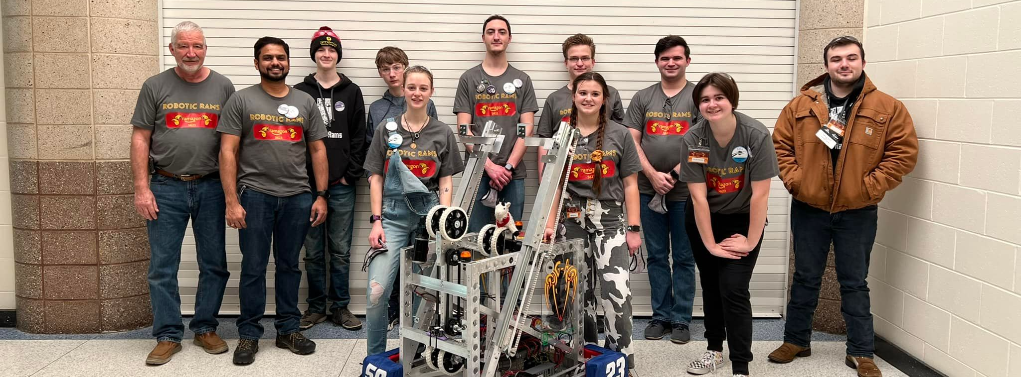 The Robotic Rams team of High School students standing behind the robot they made. The robot has four wheels and is silver.