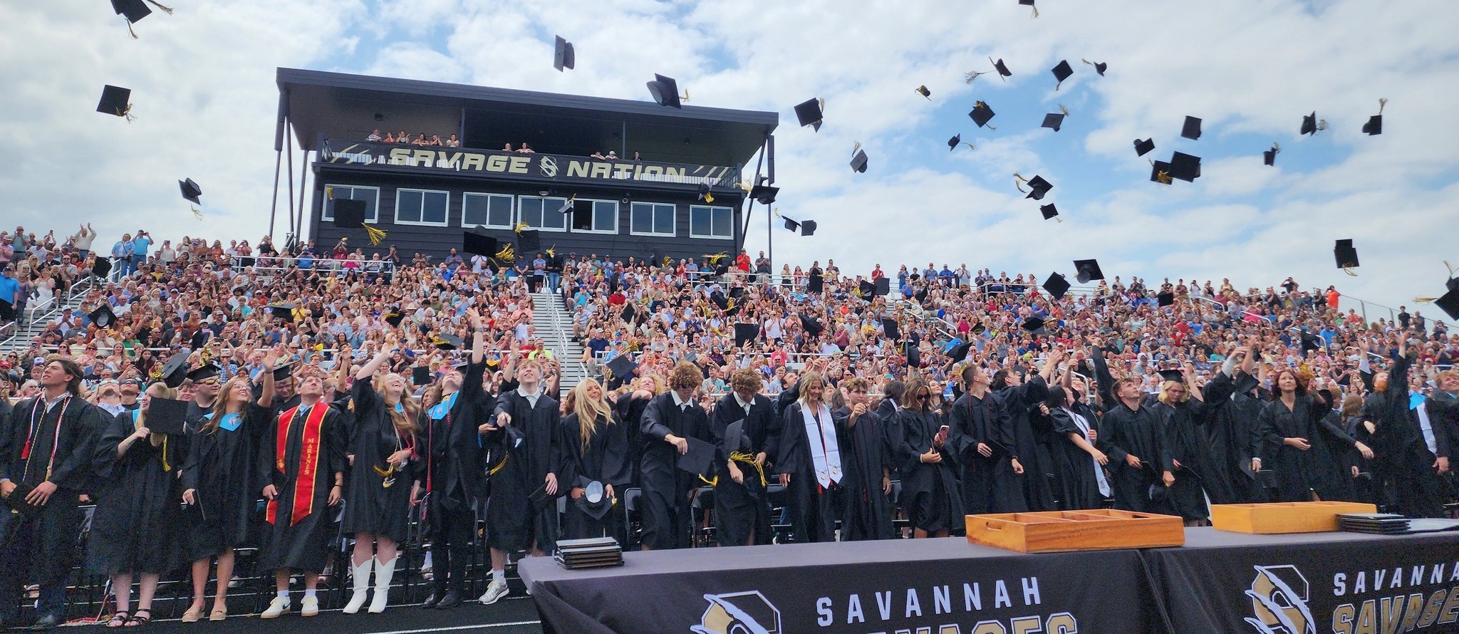 wide shot angle of graduation.  Parents are in the bleachers cheering and taking pictures.  Students are in front of the bleachers, thowing thier grauation caps in the air.  