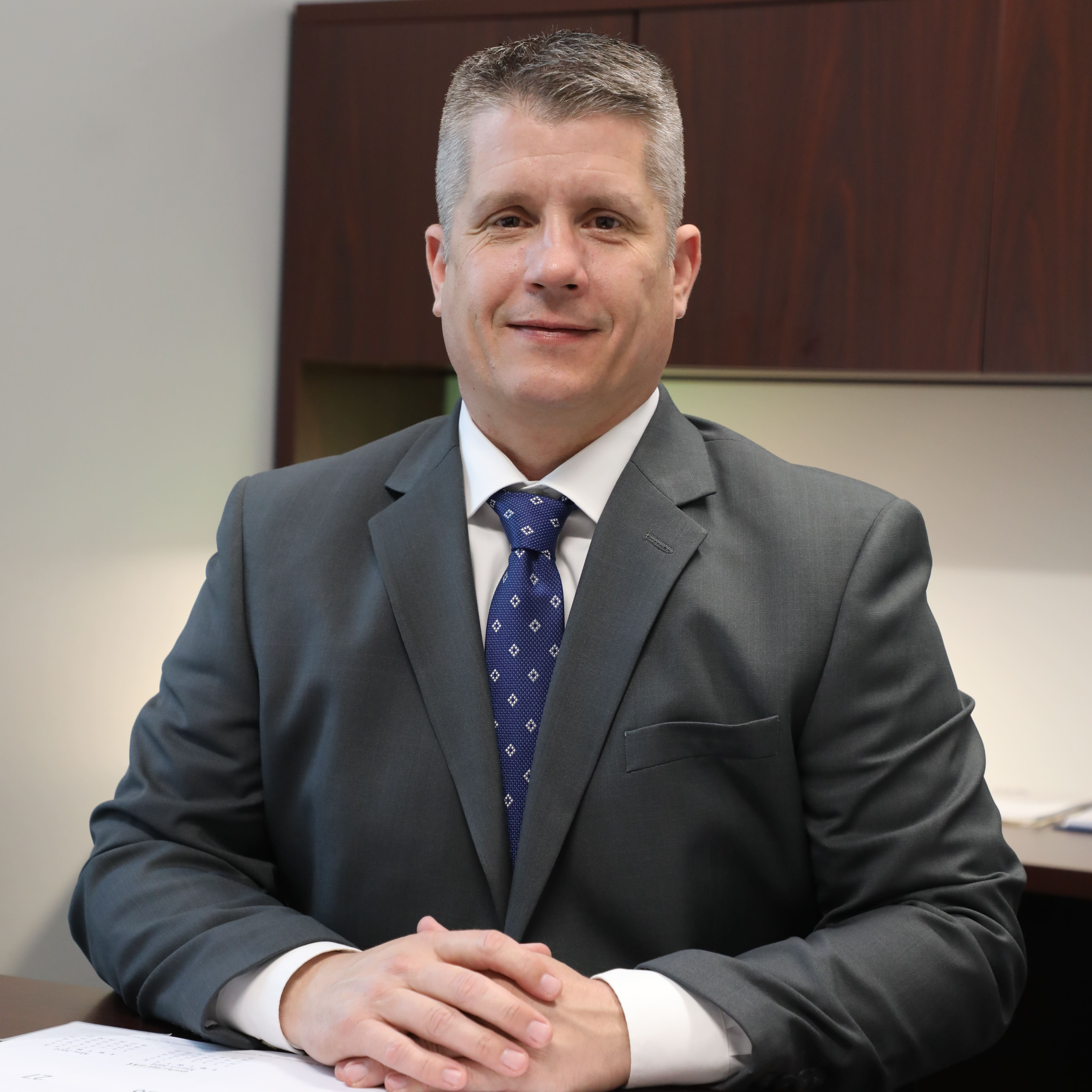 A photo of Superintendent Jason Fife, wearing a grey suit jacket, white collared shirt, and blue patterned tie, sitting at a desk with his hands clasped in front of him