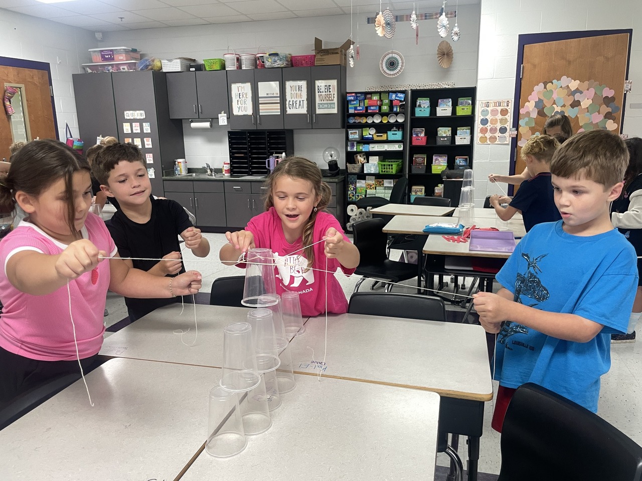 elementary students using a rubberband tied with strings to move a plastic cup