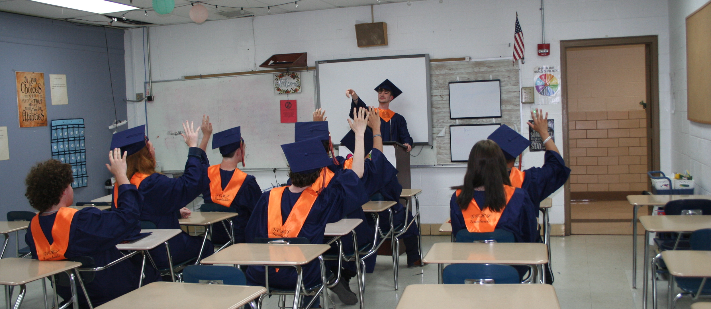 Students seated at desks in classroom, one student in front at podium, all in blues and orange caps and gowns