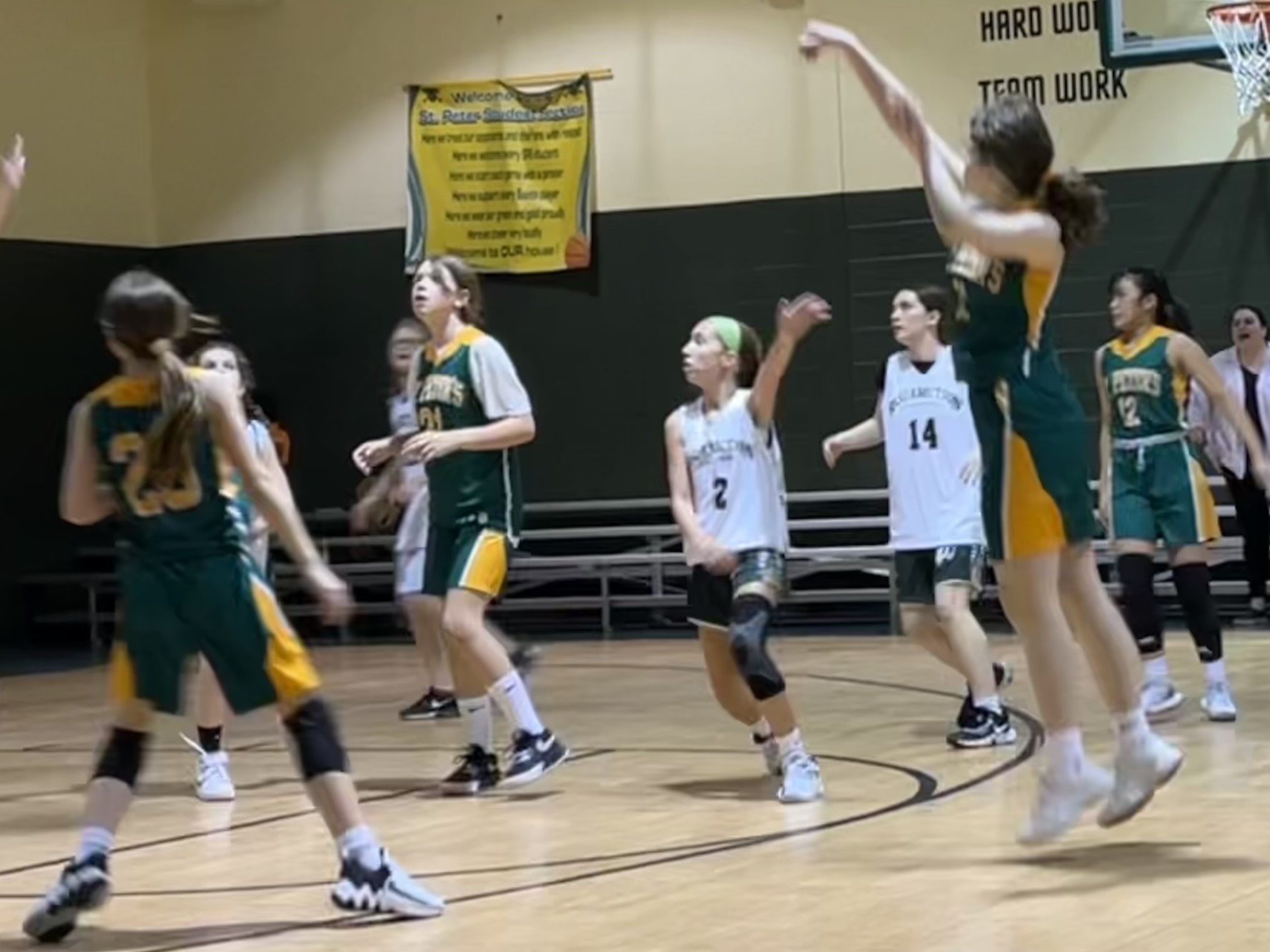 A middle school student wearing a green and gold uniform jumps to shoot a basketball.