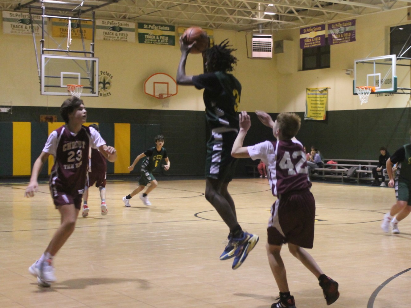 A St. Peter student wearing green and gold uniform taking a basketball jump shot against two opposing team members wearing maroon.