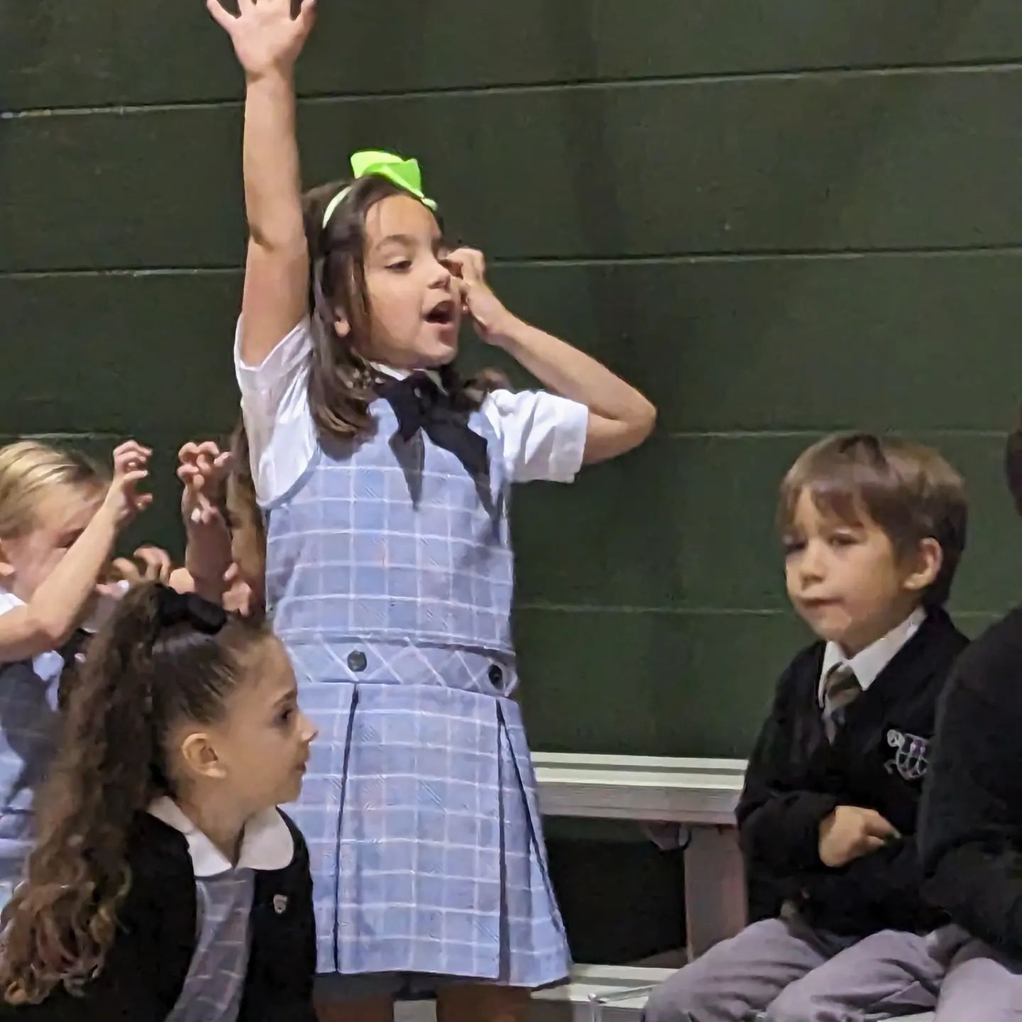a kindergarten student with medium skin tone, straight brown hair and a bow, wearing a gray St. Peter School jumper, raising her hand