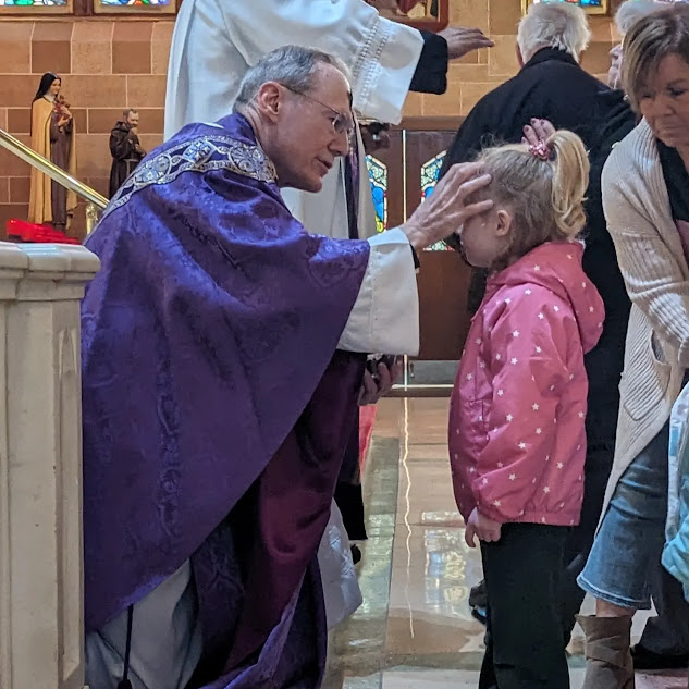 A girl in a pink coat receiving ashes for Ash Wednesday from Fr. Tim Byerley, wearing purple vestments