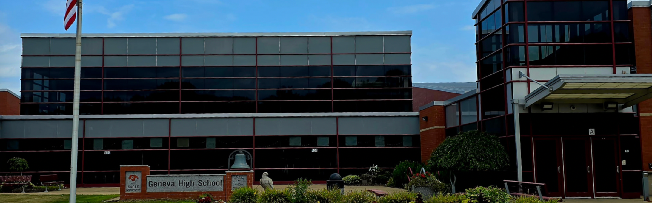 A school building with glass windows and an American flag on a pole. A grassy lawn and shrubs are in front.