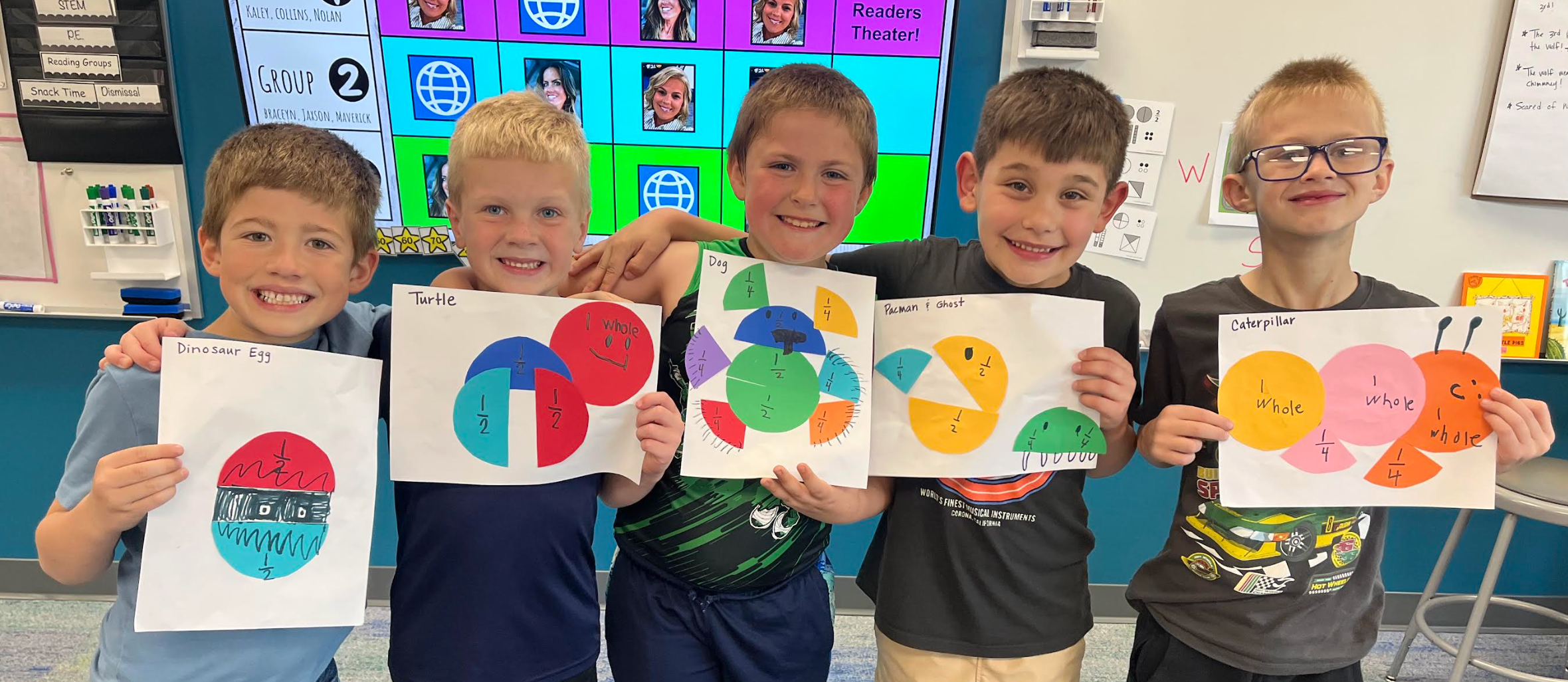 Five boys stand in a classroom, holding colorful circle diagrams. The wall behind them features a digital board displaying various images and text.