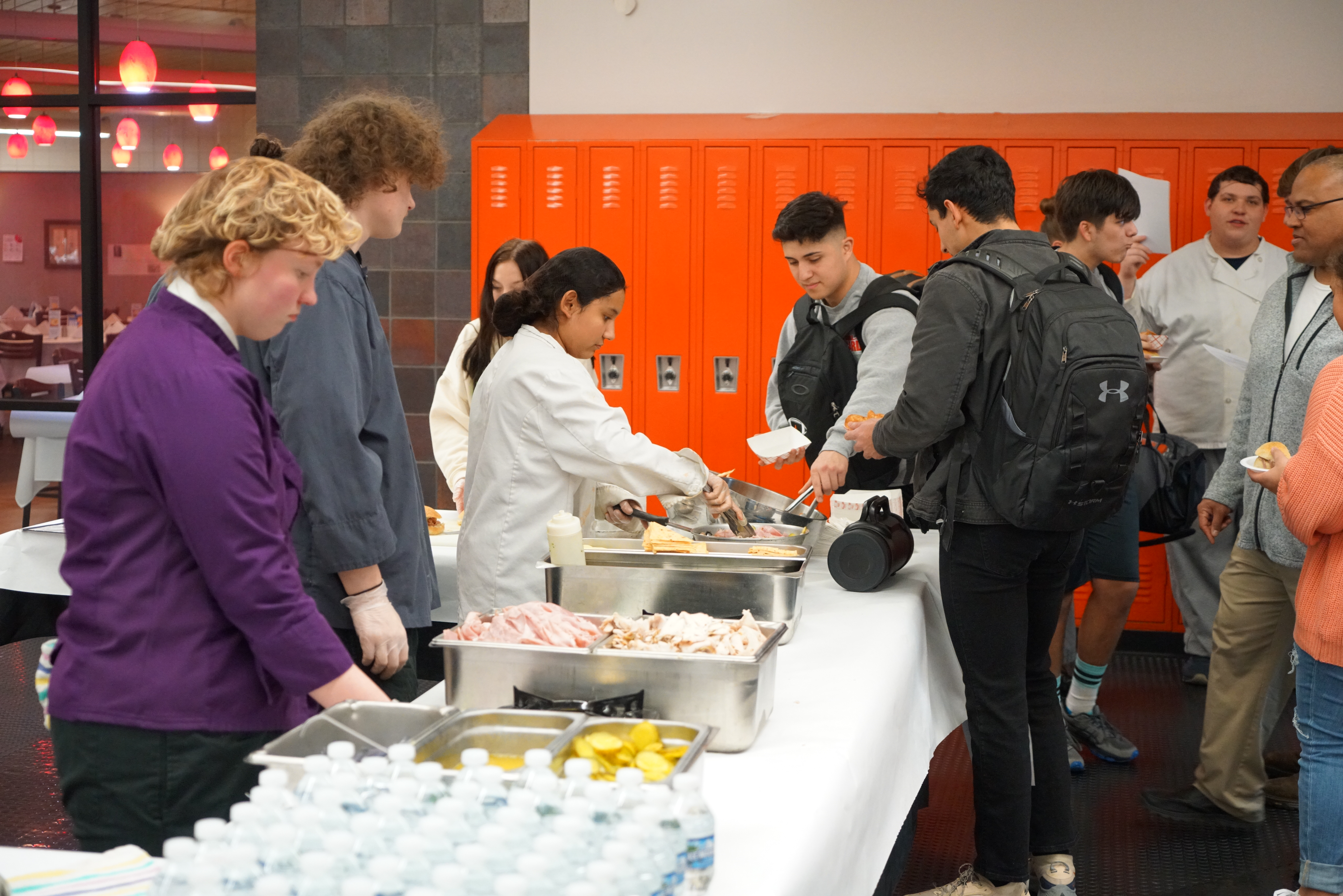 students in hair nets serve other students on a buffet line in front of orange lockers
