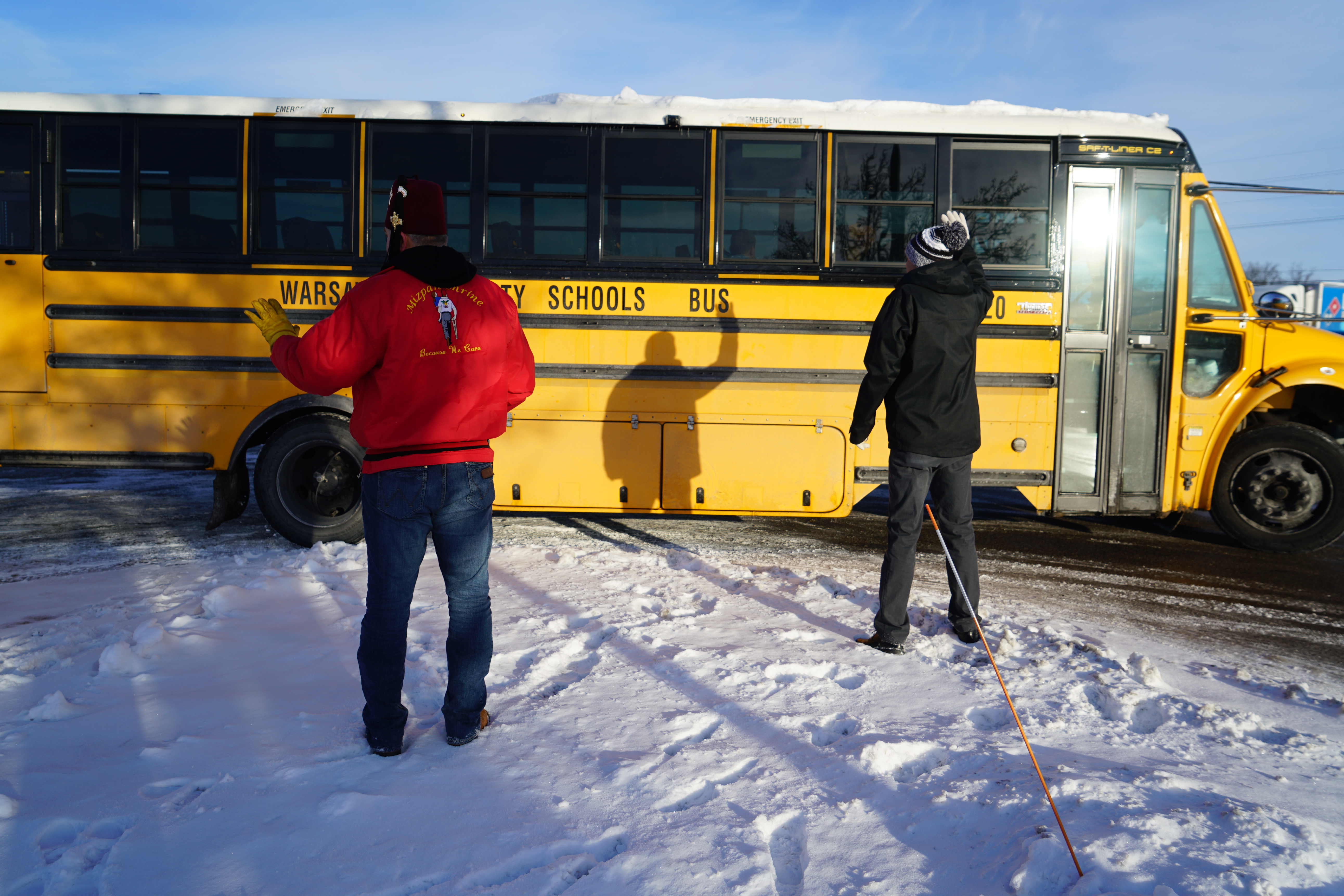 Two adults stand and wave at a warsaw school bus in the snow
