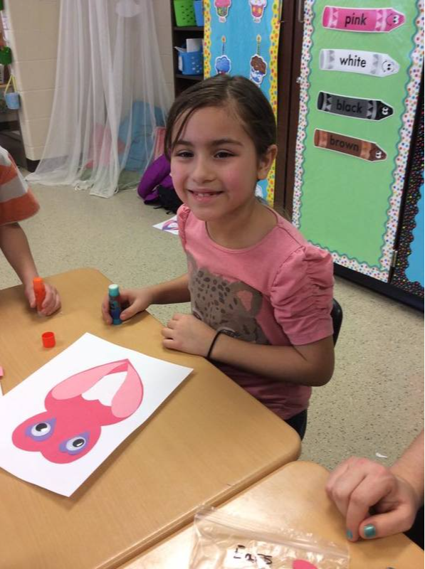 A young girl smiles holding a glue stick and working on her pink craft project