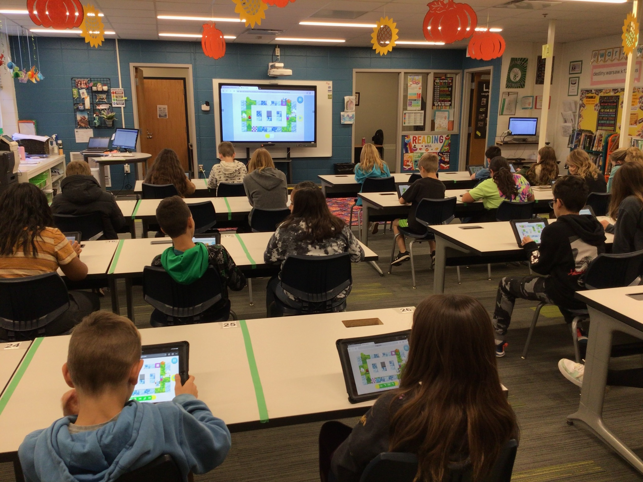 Students in a classroom on iPads all playing the same game that is projected on the screen at the front of the room