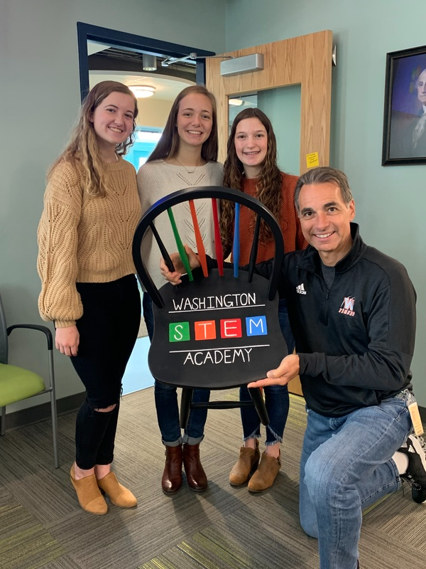 Three students with a teacher holding a chair with writing on the seat that reads: Washington STEM Academy