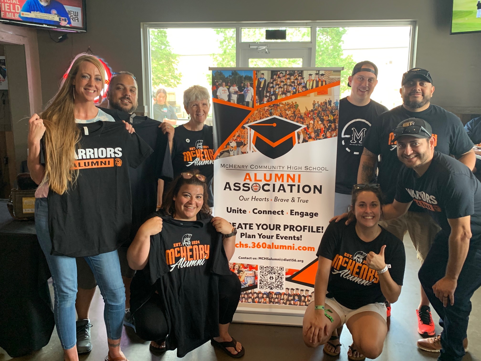 Alumni holding or wearing McHenry Alumni shirts in front of Alumni association banner