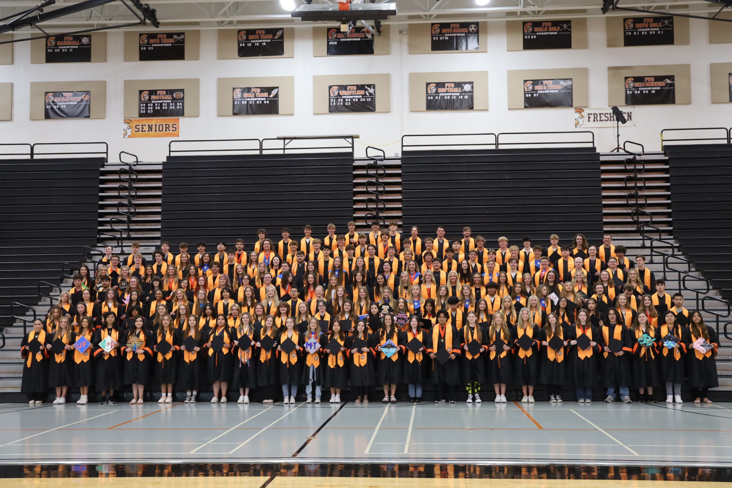 Distinguished Warrior graduates 2024 in caps and gowns with orange Distinguished Warrior stoles posing in front of bleachers 