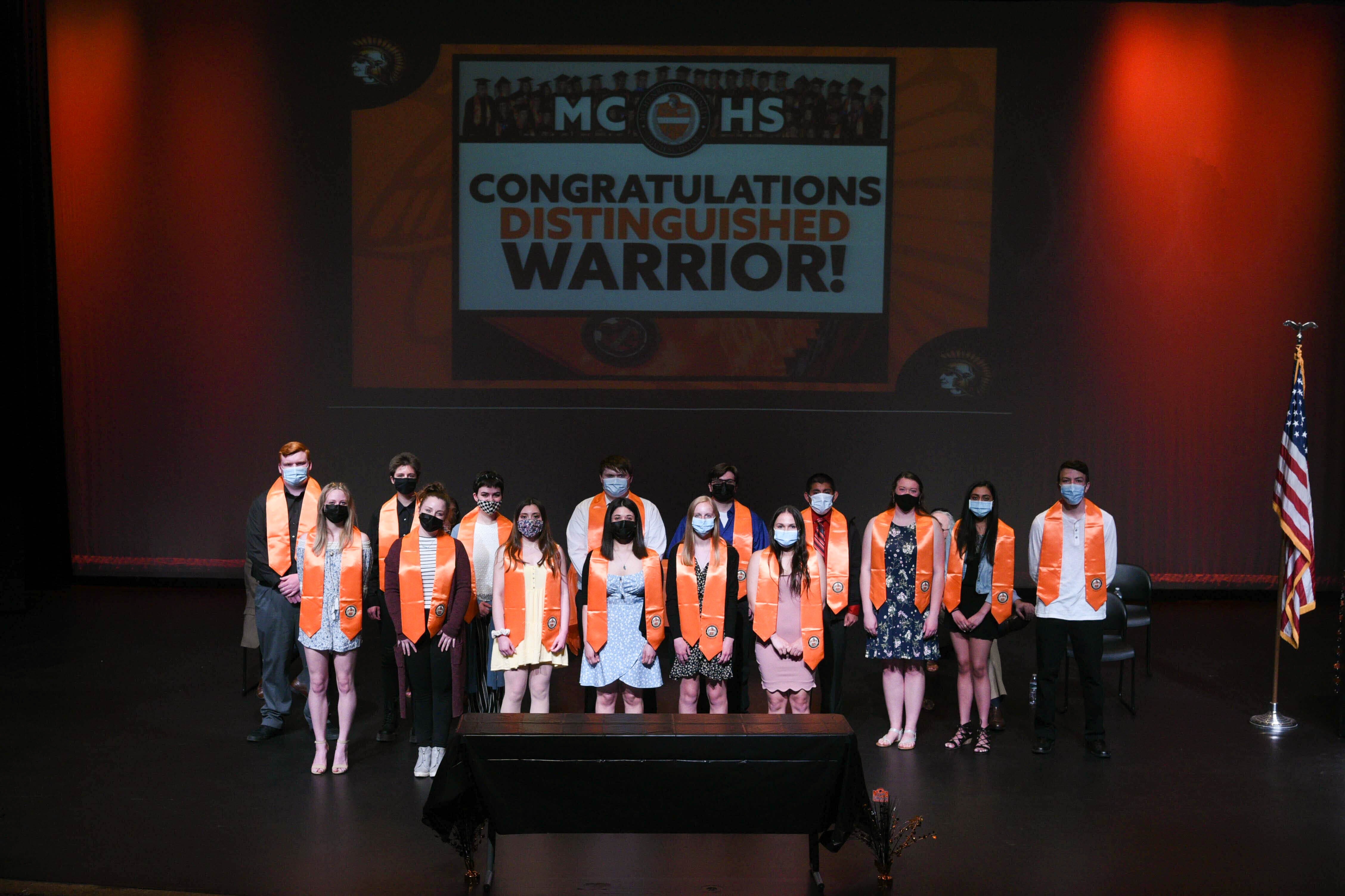 Distinguished Warrior smaller group wearing stoles on stage with masks. Congratulations Distinguished Warrior slide in the background and American flag next to them. Class of 2021