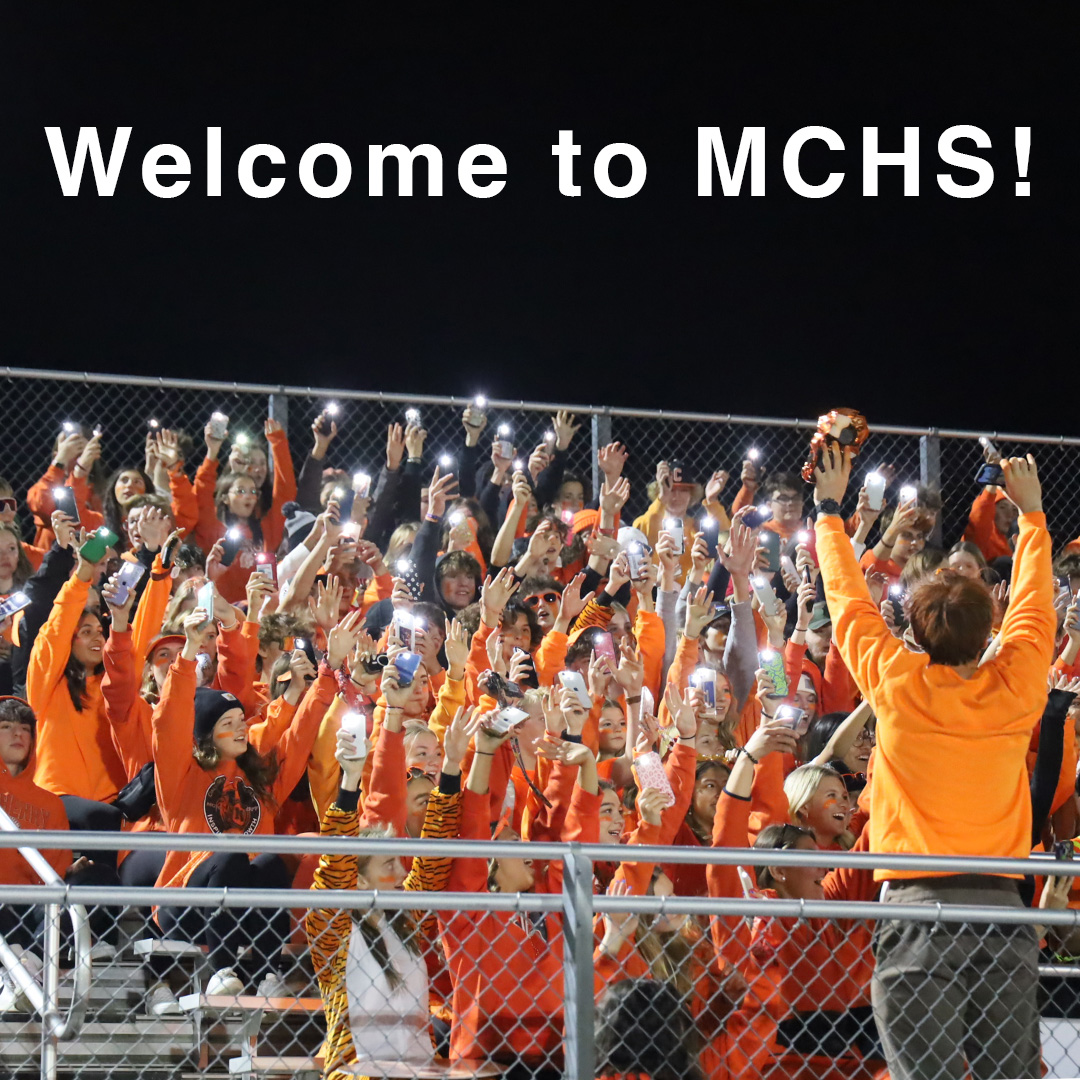 Student in football stands wearing orange waving their flashlights on their phones. One person leading them