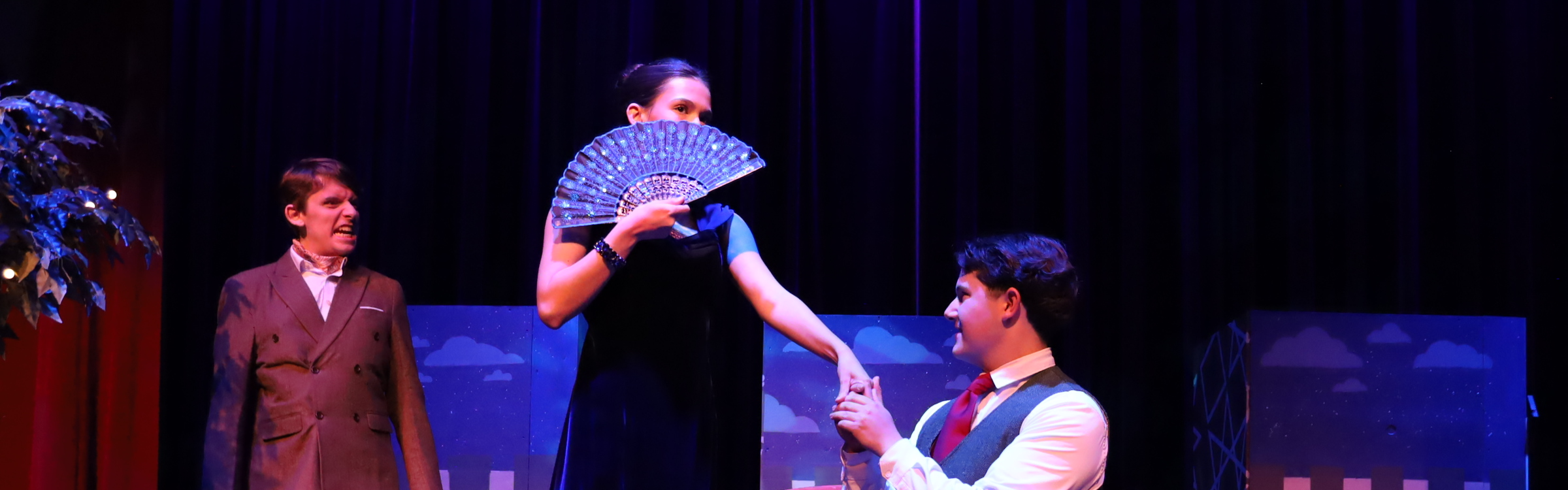 Student actress with fan over her face other student on his knees holding her hand. Student actor in the background angrily looking at them