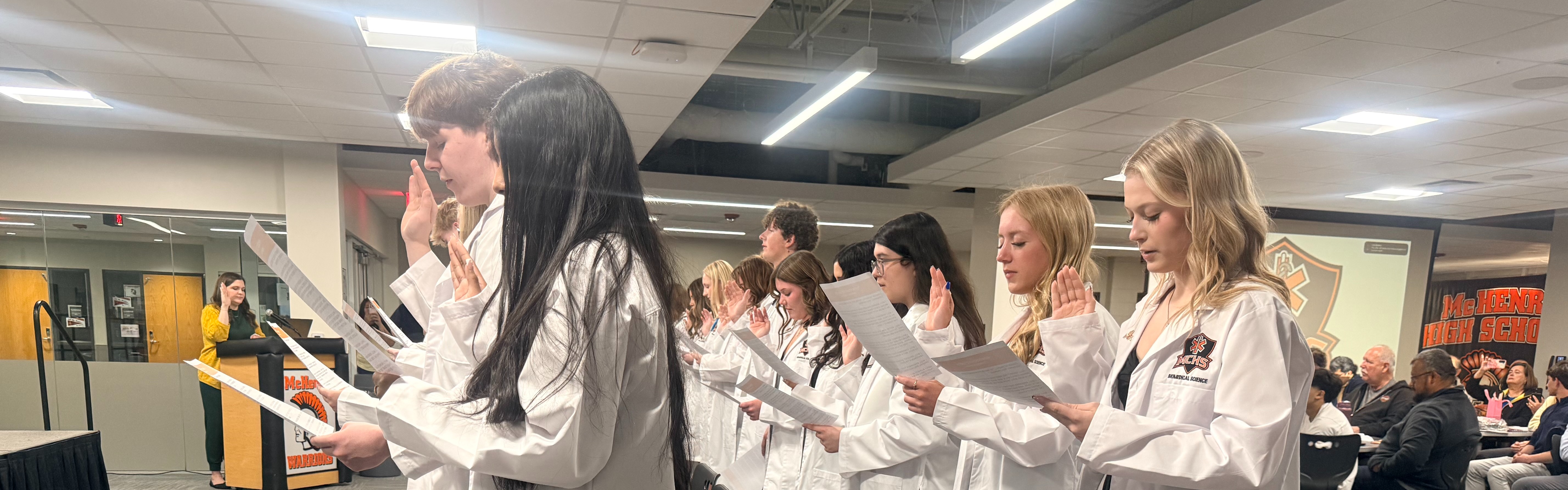 Students in white coats standing up holding hands up to do an oath