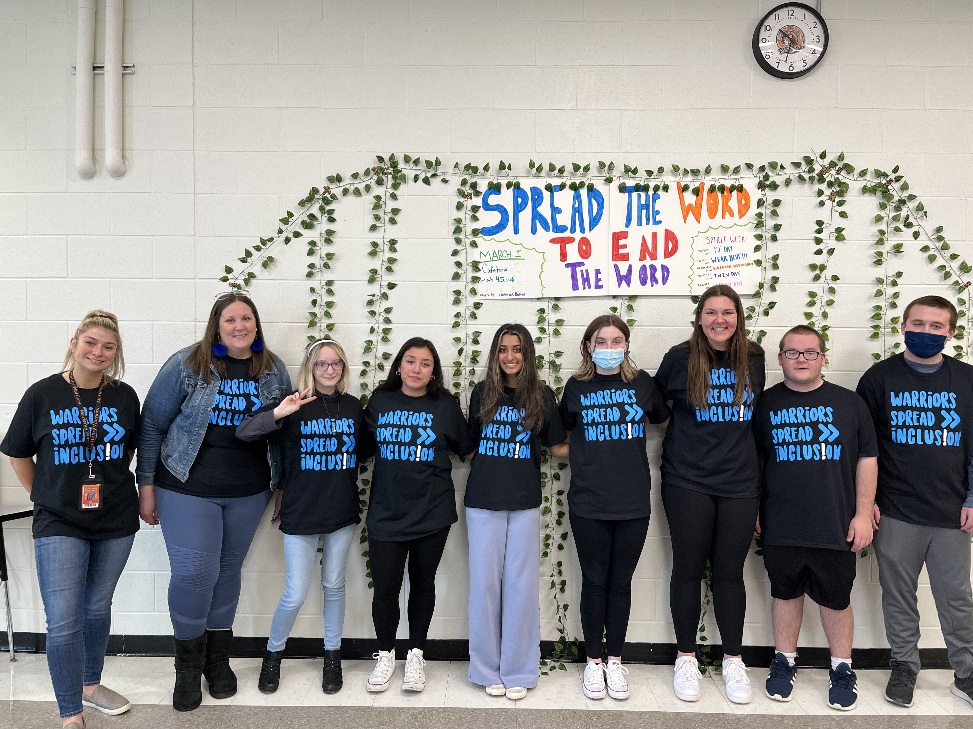 Students and staff stand against a white wall decorated with artificial leafy vines and a poster that reads "Spread the word to end the word: Warriors Spread Inclusion"