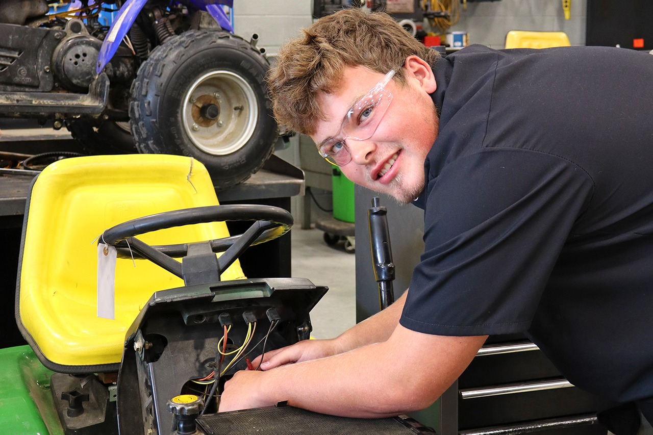 Man wearing glasses and a black shirt working on a mower in a shop with a yellow four-wheeled vehicle behind.