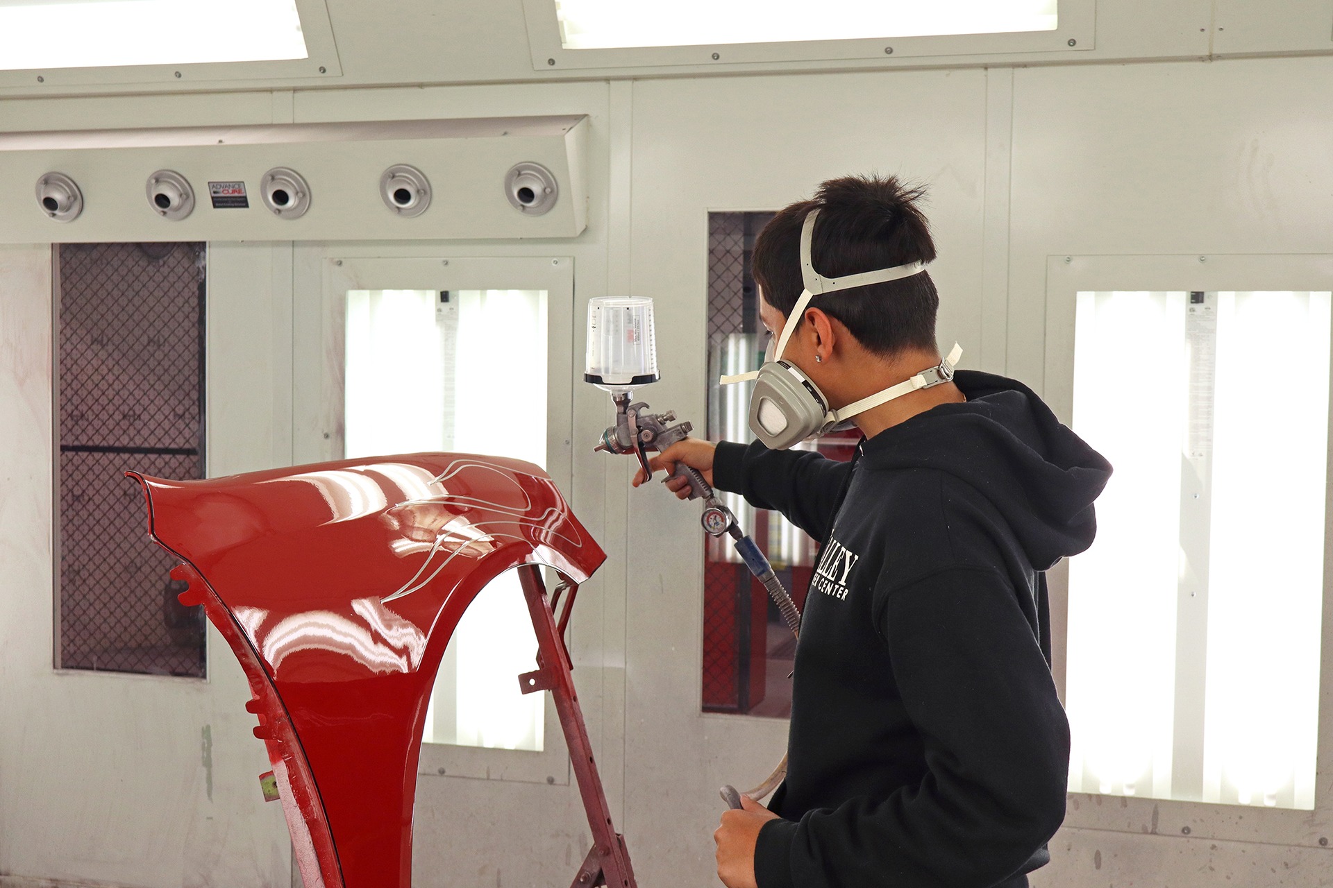 A person uses a paint sprayer to paint a red car part in a white, windowed room.