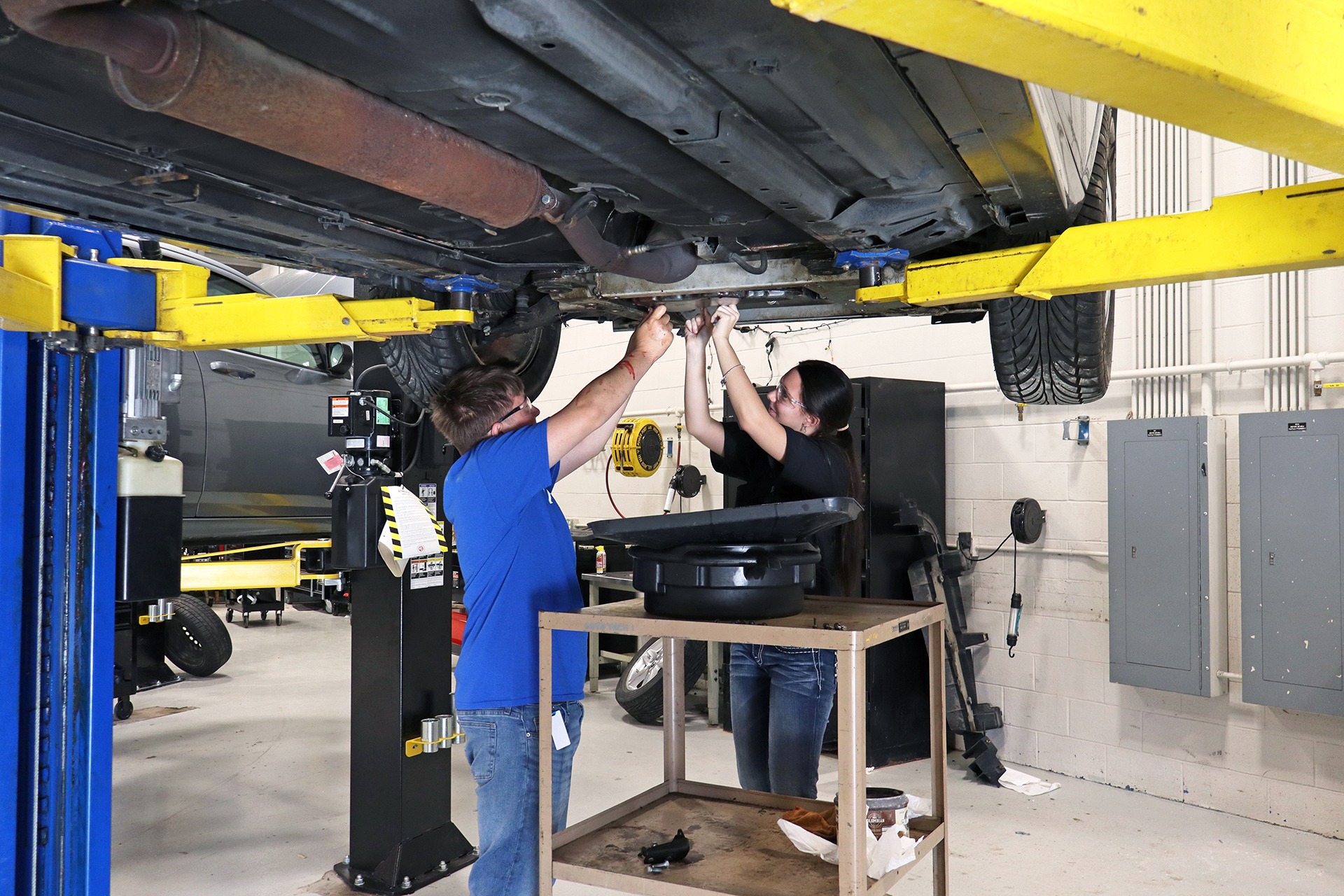 Two people in a garage working underneath a lifted car. One wears a blue shirt and the other a black shirt.