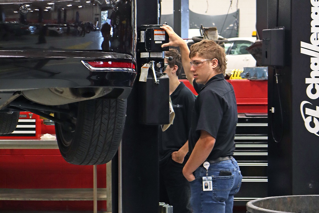 A man in a shop lifts a black car on a jack. Another man stands nearby, and a large metal part is visible in the foreground.