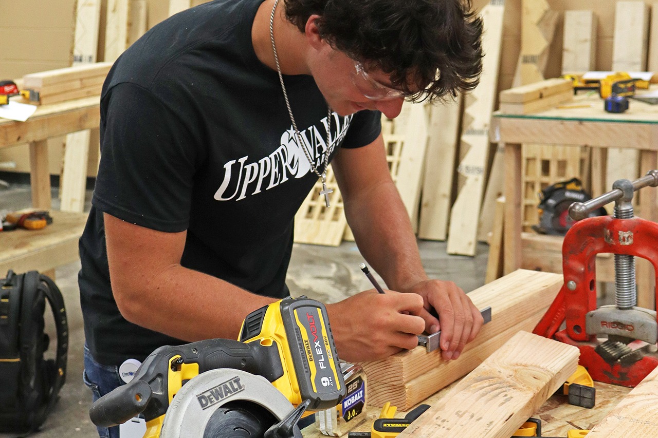 A person cuts a plank with a saw in a workshop, with wooden materials and tools around.