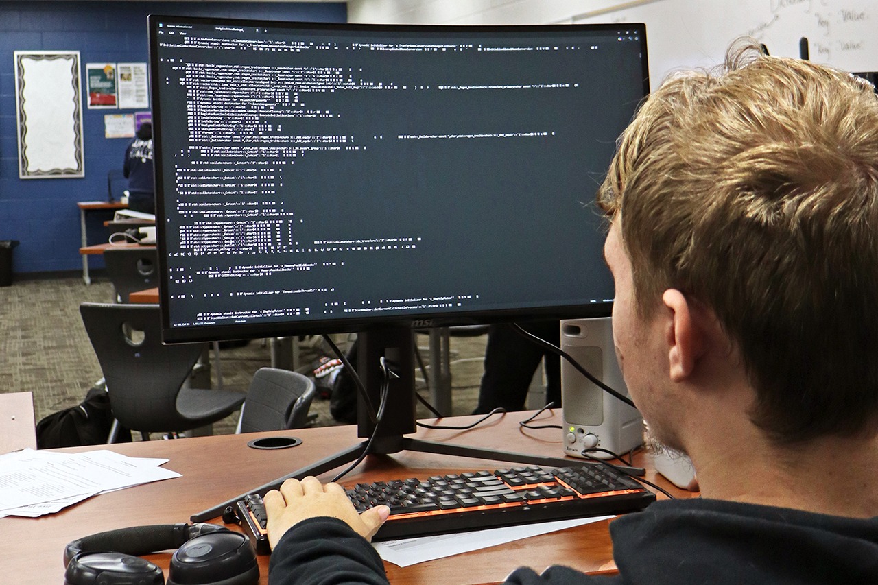 A person types on a keyboard in front of a monitor with a code interface displayed.