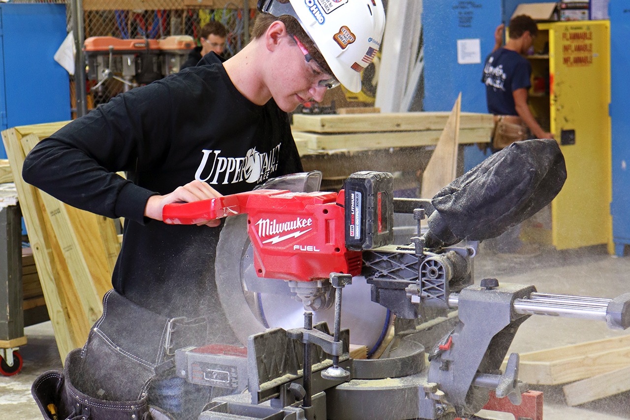 A person wearing a white helmet and black long-sleeve shirt uses a circular saw in a workshop.