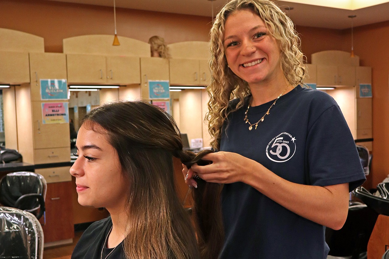 A woman sits in a chair, getting her hair done by a smiling stylist in a salon.