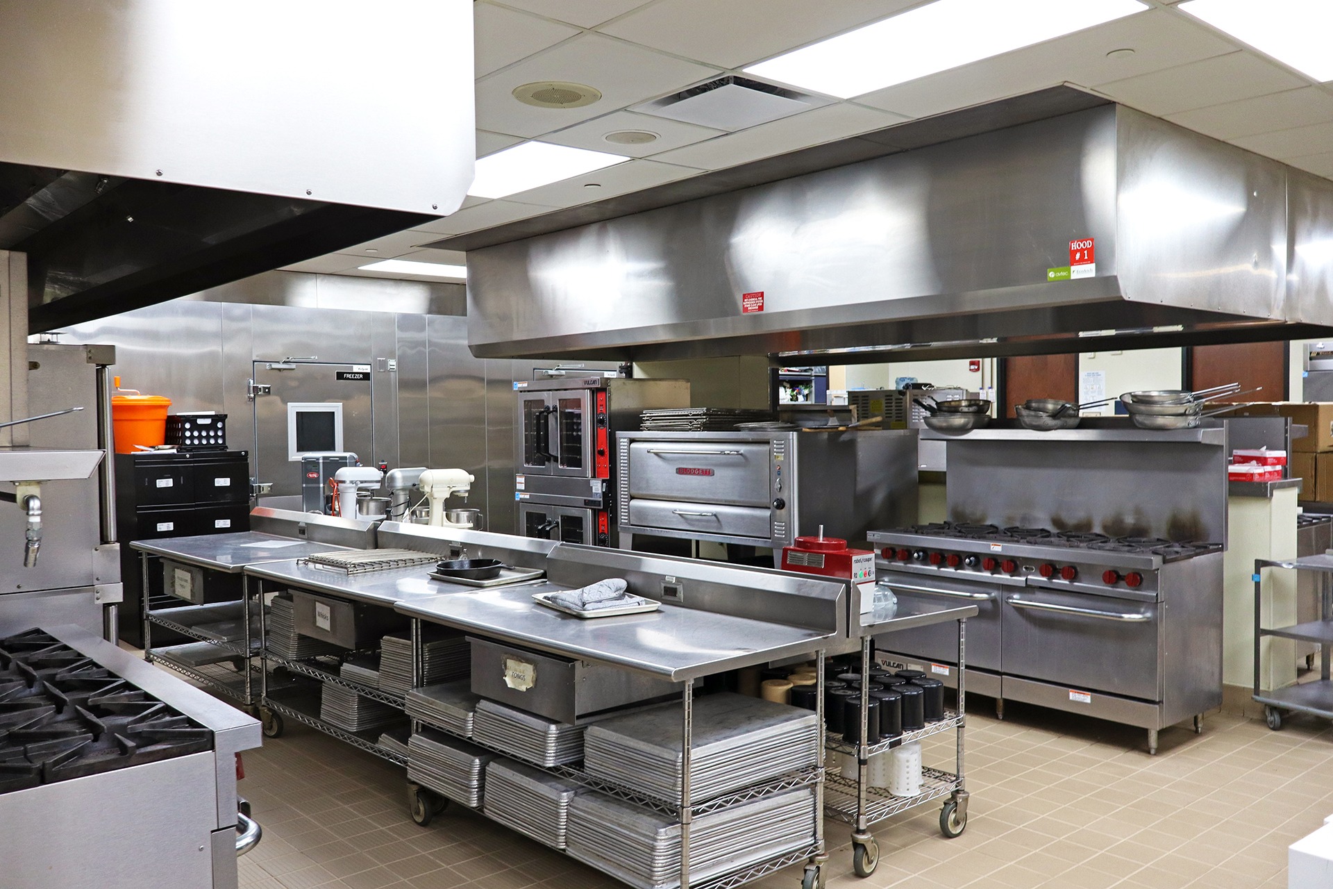An indoor commercial kitchen features a tiled floor, stainless steel appliances, and overhead lighting.
