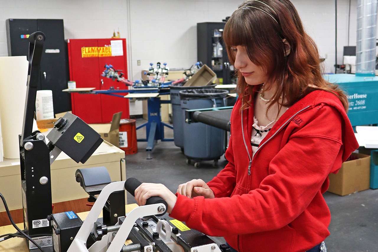 A woman in a red jacket operates a machine on a table in a factory-like setting.