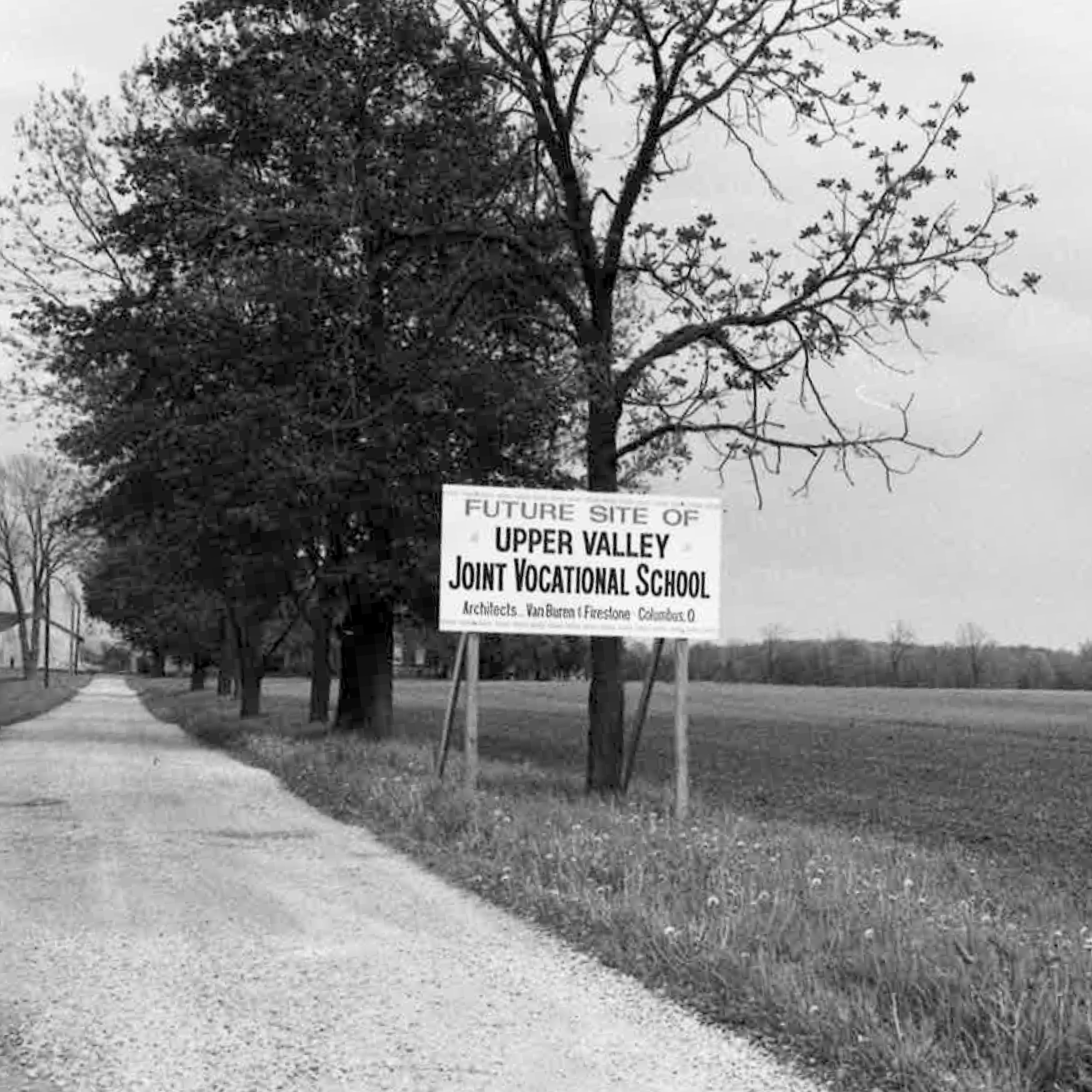 A black-and-white photo of a dirt road flanked by trees. A sign reads "Future Site of Upper Valley Joint Vocational School."